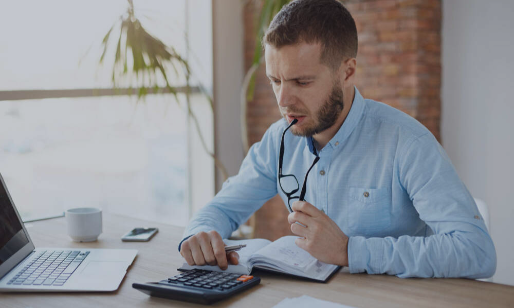 Hombre trabajando como autónomo en su escritorio, utilizando una calculadora y revisando un cuaderno mientras piensa.