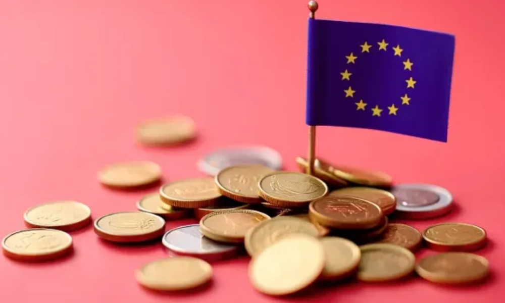 Montón de monedas europeas junto a una pequeña bandera de la Unión Europea sobre fondo rojo, simbolizando la fiscalidad y los impuestos en el ámbito comunitario.