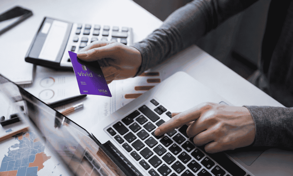 Persona sosteniendo una tarjeta Visa de Vivid morada mientras utiliza un ordenador portátil.