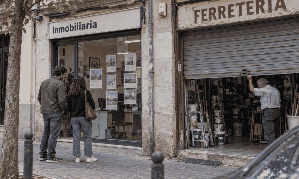 Dos personas observan el escaparate de una inmobiliaria mientras un ferretero baja la persiana del local contiguo.